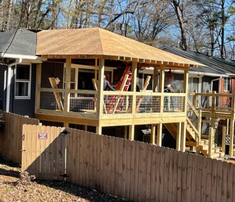 Backyard view of a brick house with a newly framed wooden deck under construction and visible workers on site.