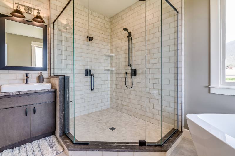 Modern glass-enclosed walk-in shower with white subway tiles, black fixtures, and a black-framed shower wand. At left: dark vanity with sink and mirror.